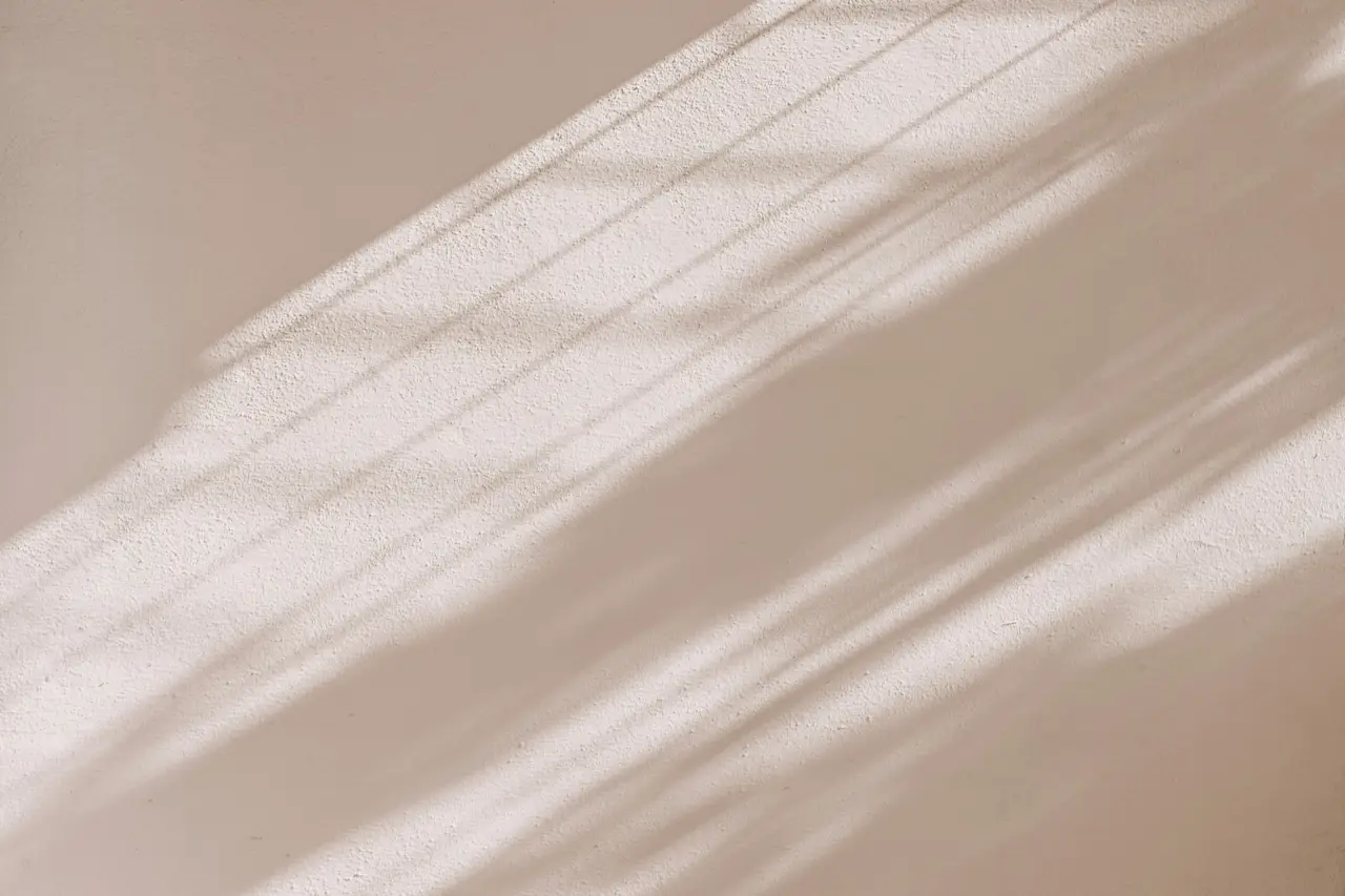Mur de séjour enduit à la chaux, sable clair, traversé en diagonale par la lumière rasante du matin filtrée par un store : le grain de l’enduit apparaît dans chaque bande de lumière